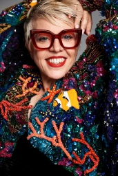 Close-up portrait of a smiling woman with short silver hair wearing red-framed glasses and a heavily embellished sequined jacket.