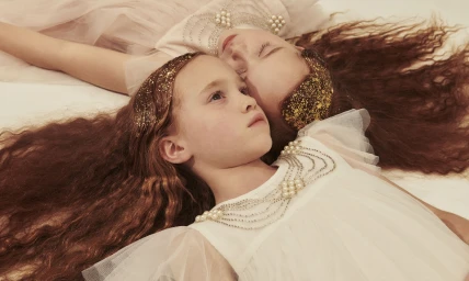 Two young girls with long reddish-brown hair lying down wearing white dresses and beaded headpieces.