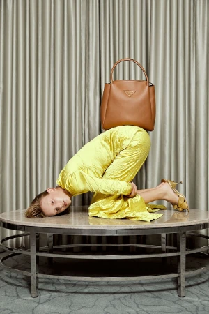 Model in a yellow dress kneeling on a round coffee table with a brown leather handbag balanced on their back