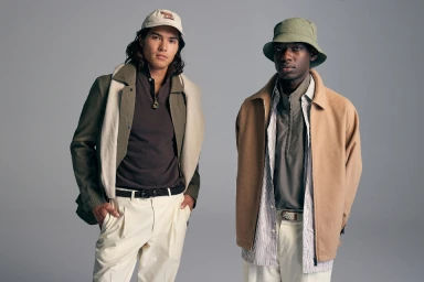 Two male models posing against a grey studio backdrop wearing layered casual outerwear and hats