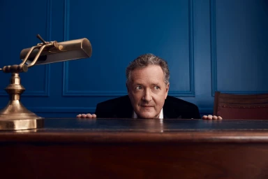 Piers Morgan peeking over a wooden desk with a brass desk lamp in a room with blue paneled walls