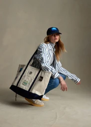 Woman in blue cap and striped shirt crouching with a large canvas tote bag against a neutral studio backdrop
