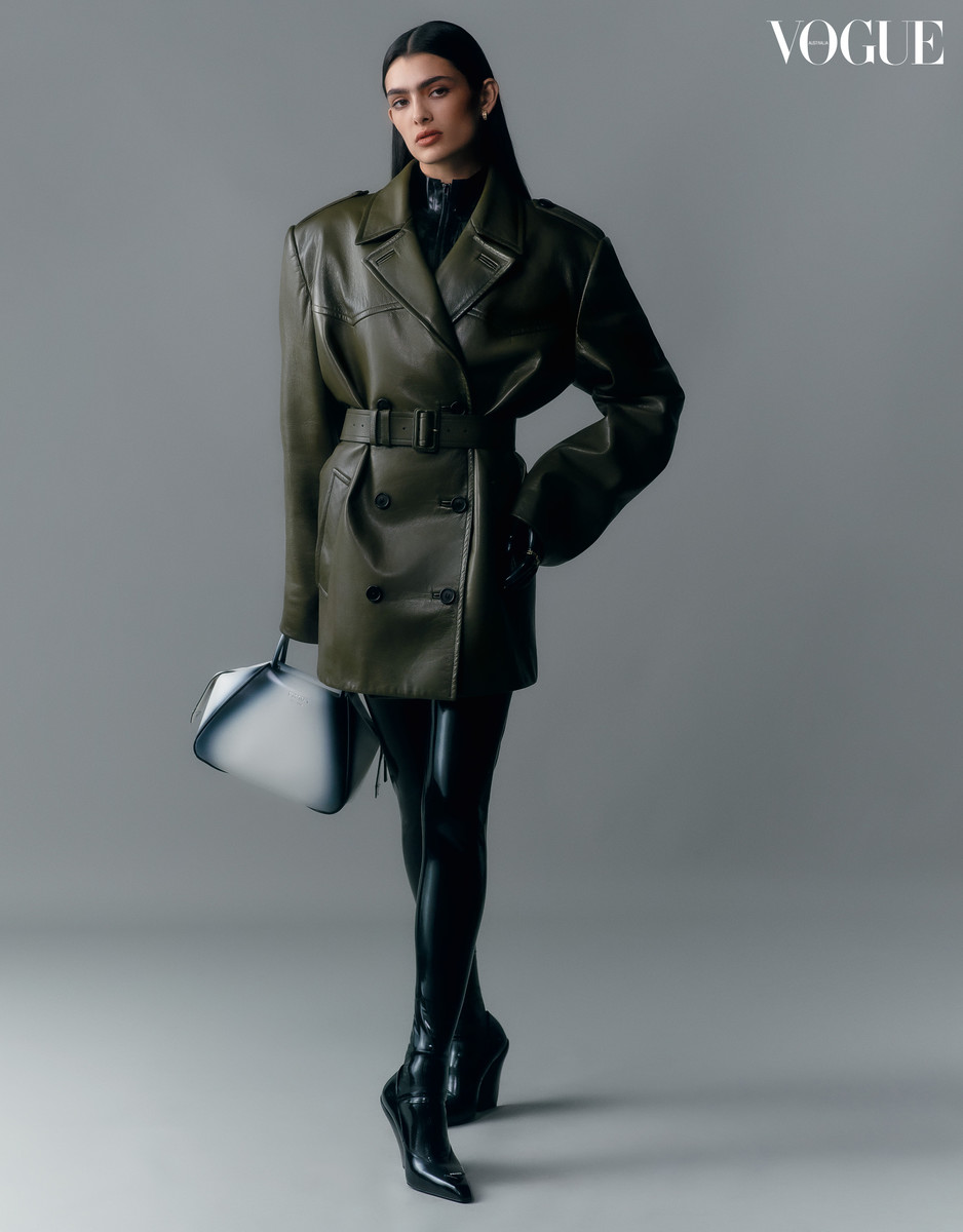 A woman in a green leather coat and black boots poses with a white handbag against a grey backdrop in a fashion editorial shot.