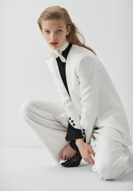 Fashion editorial shot of a crouched woman in a white suit with a black shirt and patterned heels against a white background.