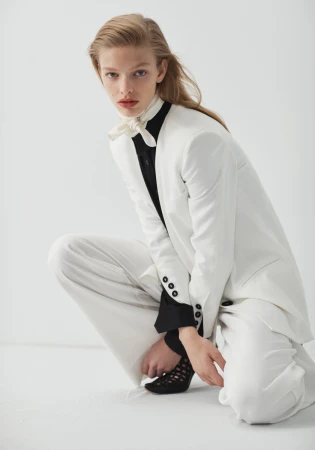 Fashion editorial shot of a crouched woman in a white suit with a black shirt and patterned heels against a white background.