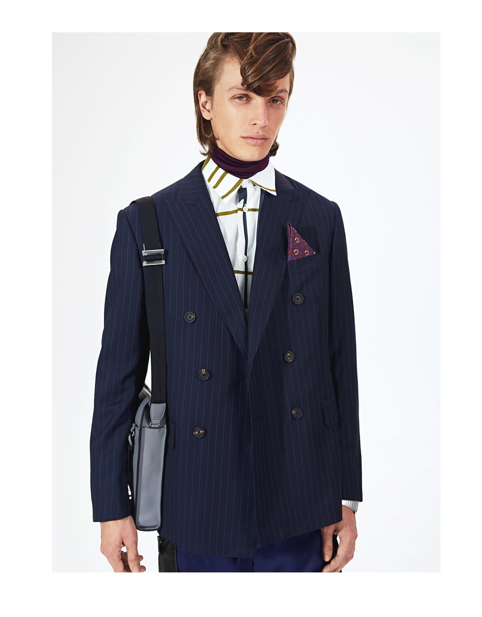 A young man poses indoors wearing a pinstripe double-breasted suit, purple turtleneck, and patterned shirt, accessorized with a shoulder bag.