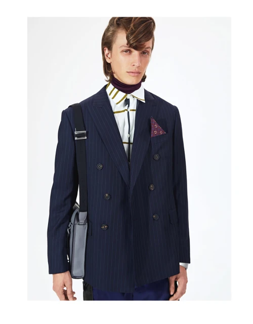 A young man poses indoors wearing a pinstripe double-breasted suit, purple turtleneck, and patterned shirt, accessorized with a shoulder bag.