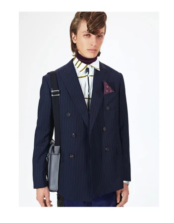 A young man poses indoors wearing a pinstripe double-breasted suit, purple turtleneck, and patterned shirt, accessorized with a shoulder bag.
