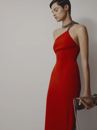 Model with short dark hair wearing a vibrant red dress with a chunky chain strap and holding a patterned handbag against a plain light background