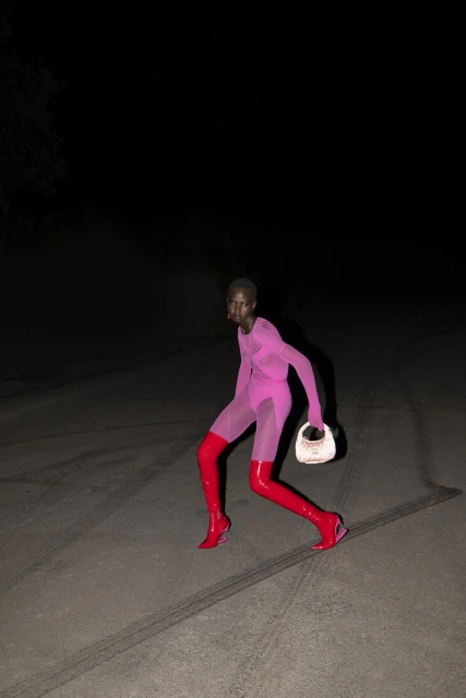 Fashion editorial shot of a Black female adult striding on an asphalt road with tire tracks at night, wearing a pink bodysuit and red boots.