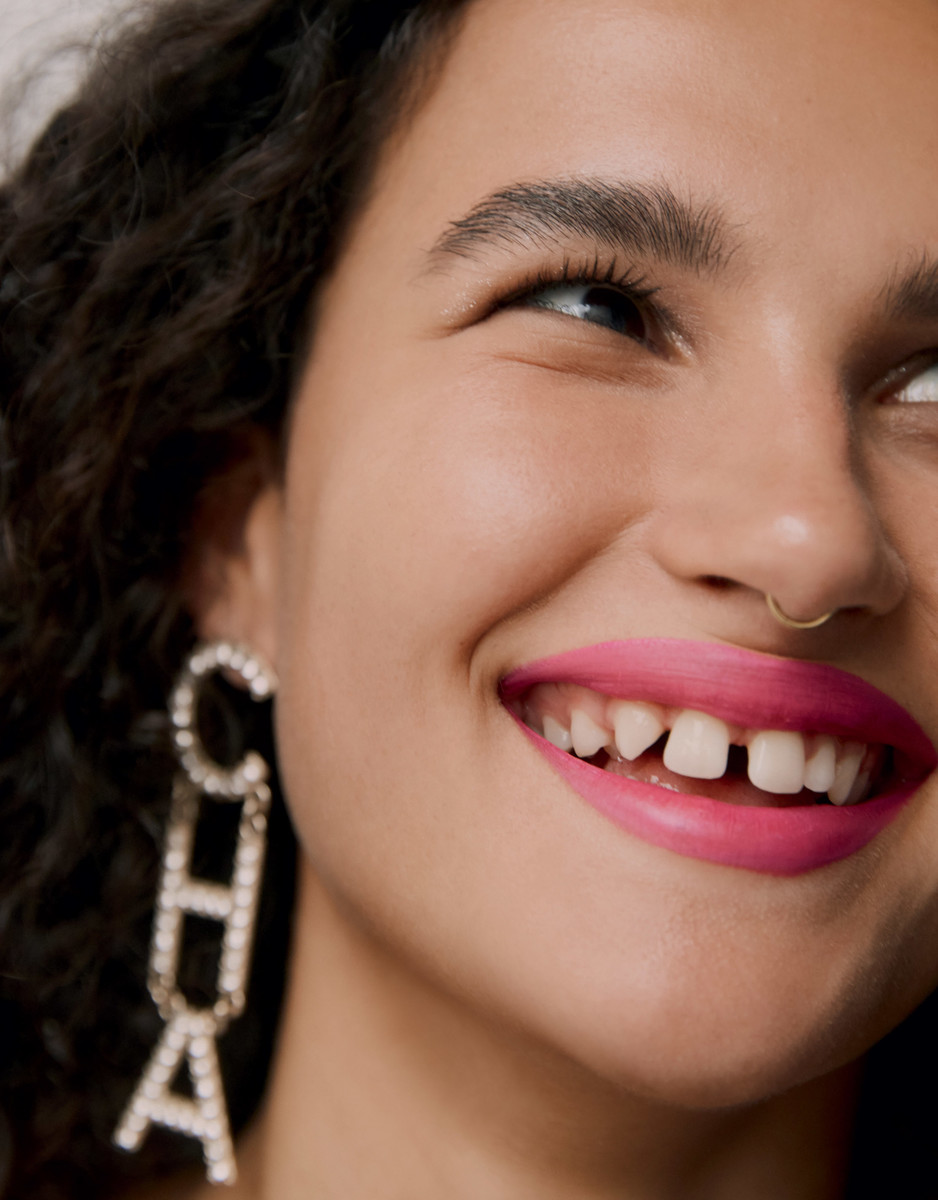Close-up portrait of a smiling woman with dark curly hair, pink lipstick, gold nose ring, and rhinestone "CHA" letter earrings.