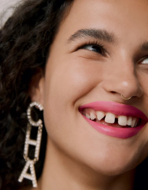 Close-up portrait of a smiling woman with dark curly hair, pink lipstick, gold nose ring, and rhinestone "CHA" letter earrings.