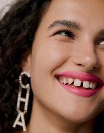 Close-up portrait of a smiling woman with dark curly hair, pink lipstick, gold nose ring, and rhinestone "CHA" letter earrings.