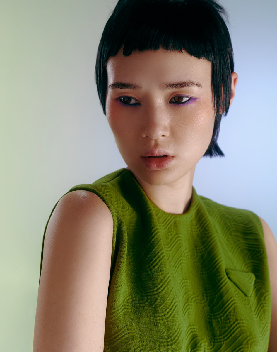 Close-up of a young Asian woman with distinctive short bangs, wearing a textured green top and vibrant makeup, looking off-camera.