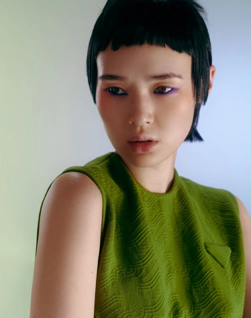 Close-up of a young Asian woman with distinctive short bangs, wearing a textured green top and vibrant makeup, looking off-camera.