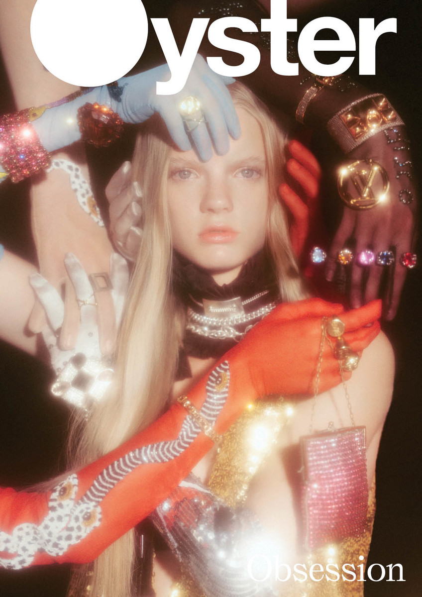 Magazine cover featuring a blonde model with several hands adorned with jewelry, surrounding her face and body against a dark background.
