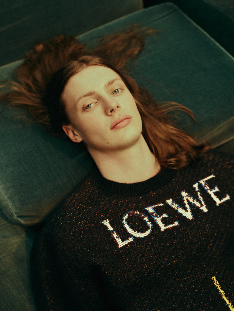 A young adult male model with long hair reclines on a green couch wearing a LOEWE logo sweater in a fashion editorial shot.