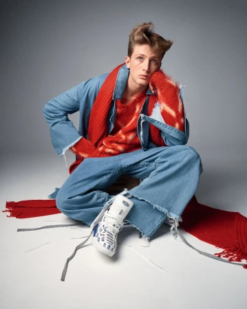 A young adult male sits cross-legged in a photo studio wearing a blue denim outfit with a red scarf and sweater and white sneakers.