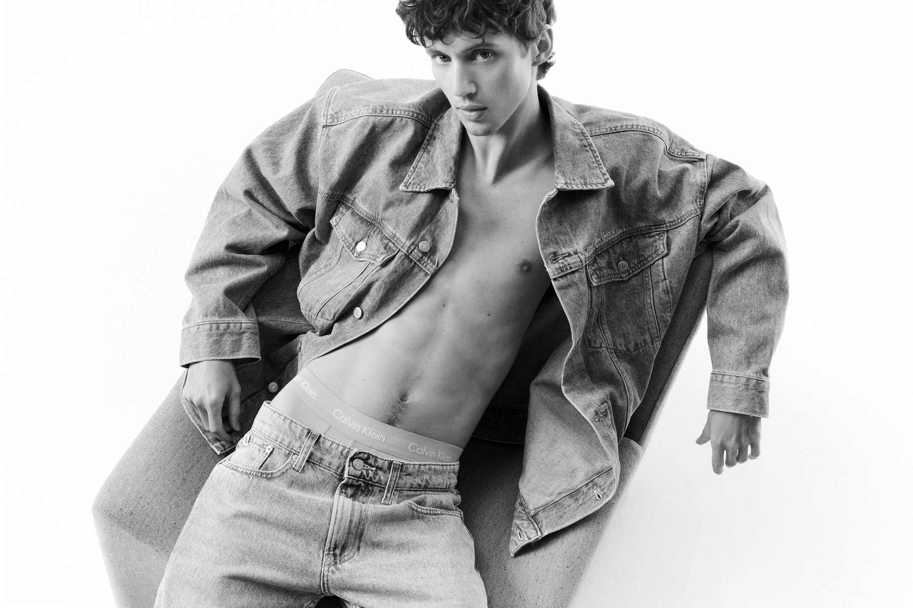 Monochrome portrait of a young male model wearing an open denim jacket and jeans seated in a studio setting