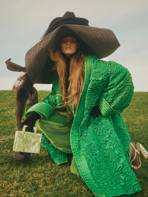 Fashion model with a large hat and green dress poses outdoors on grass, accessorized with gloves and a patterned handbag.