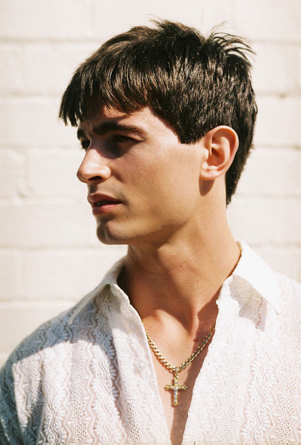 Portrait of a young adult male wearing a patterned white shirt and gold cross necklace, looking to the left against a white brick wall.