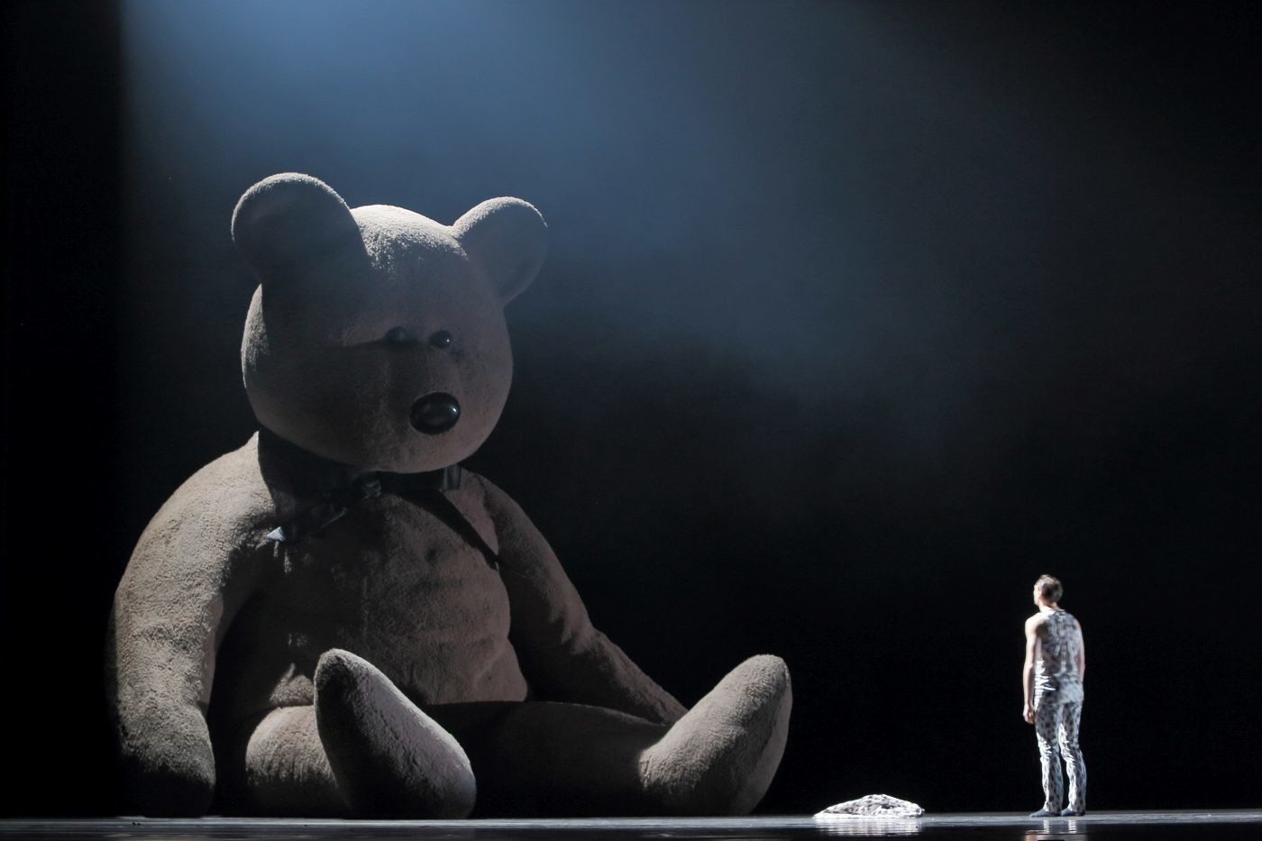 Dramatic stage lighting illuminates a giant teddy bear beside a person in a patterned full body suit on a dark stage backdrop.