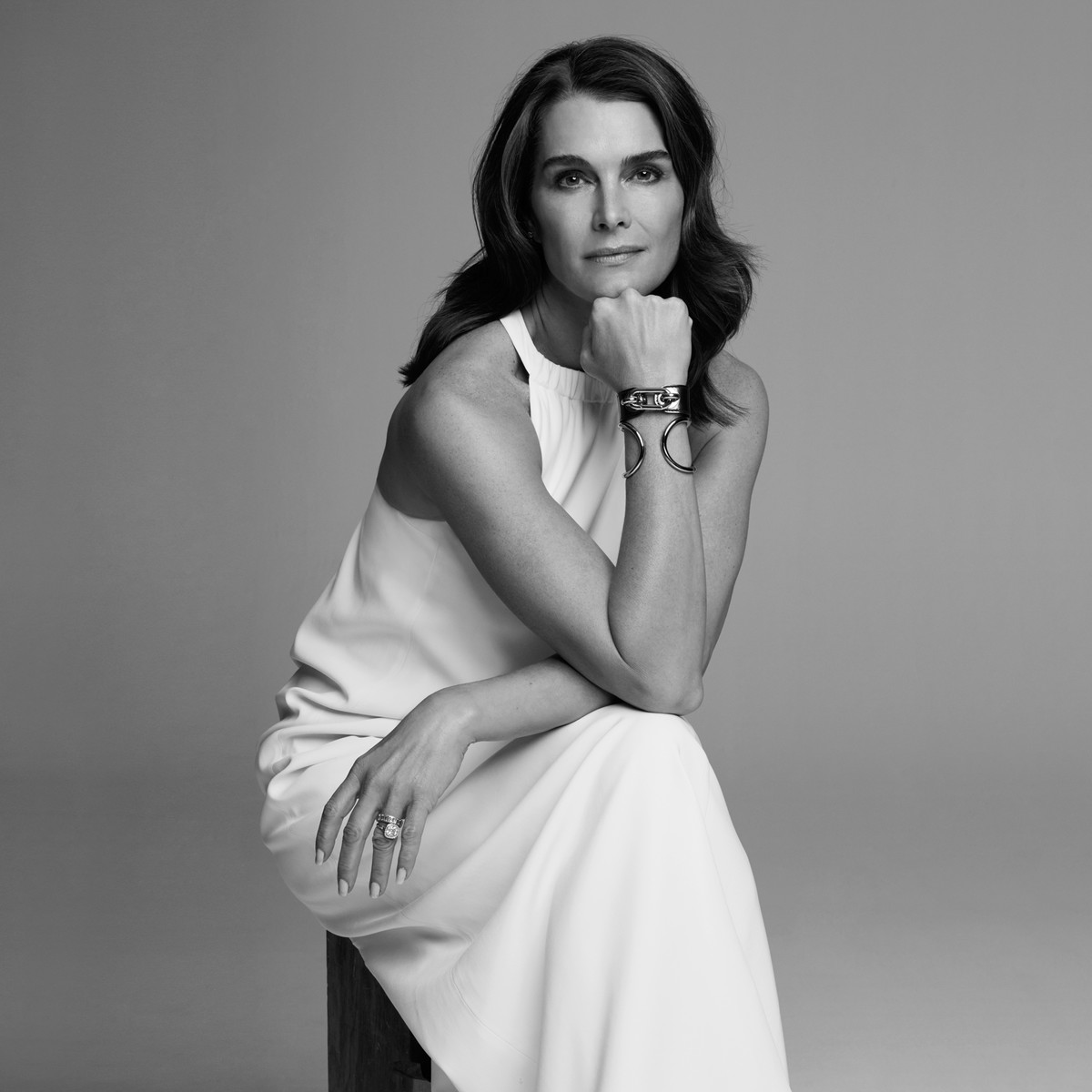 Black and white portrait of an adult female with dark hair wearing a white dress and silver bracelets, sitting in a studio setting.
