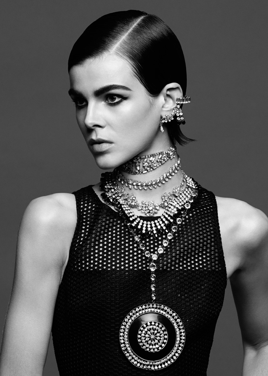 Black and white portrait of a woman wearing layers of statement jewelry, including a dramatic pendant and multiple necklaces.