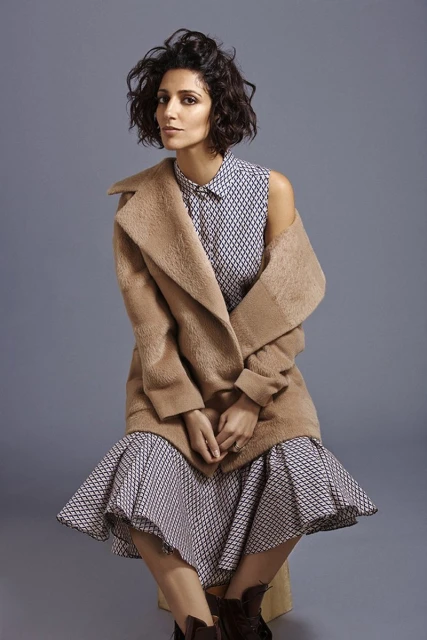A fashionable woman with curly dark hair sits while wearing a printed dress, brown coat, and matching boots against a grey backdrop.