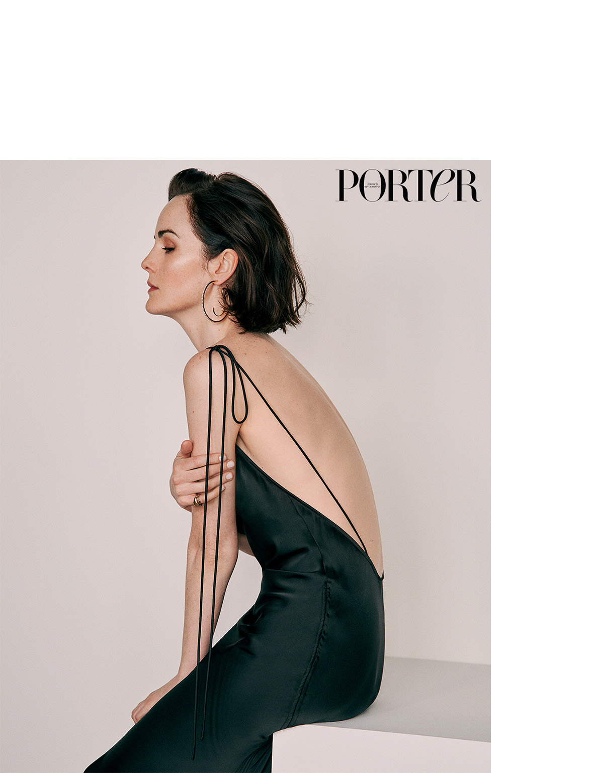 Elegant portrait of a woman in a backless black dress sitting against a plain background, showcasing beauty and fashion.