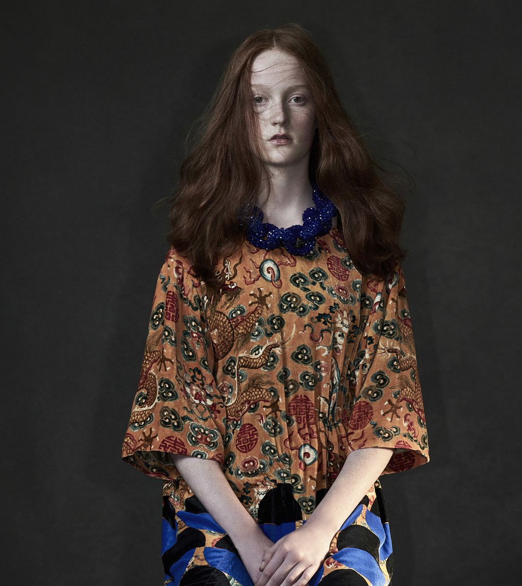 Portrait of a young woman with auburn hair wearing a patterned orange top and blue beaded necklace against a dark gray background.