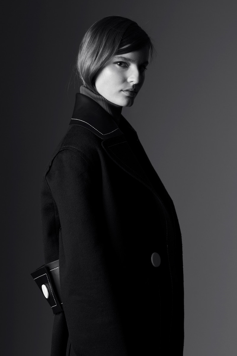 Model in a dark coat with contrasting white stitching styled by Michelle Jank against a grey background.