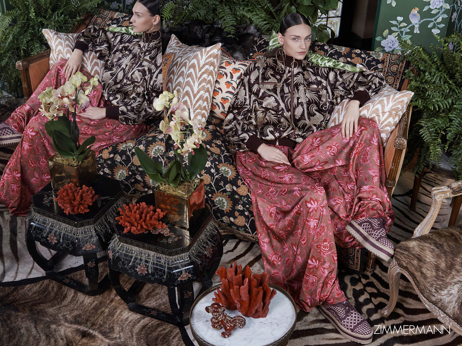 Two fair-skinned women sit on a patterned couch wearing matching printed jacket and pants outfits, with two small tables in front.