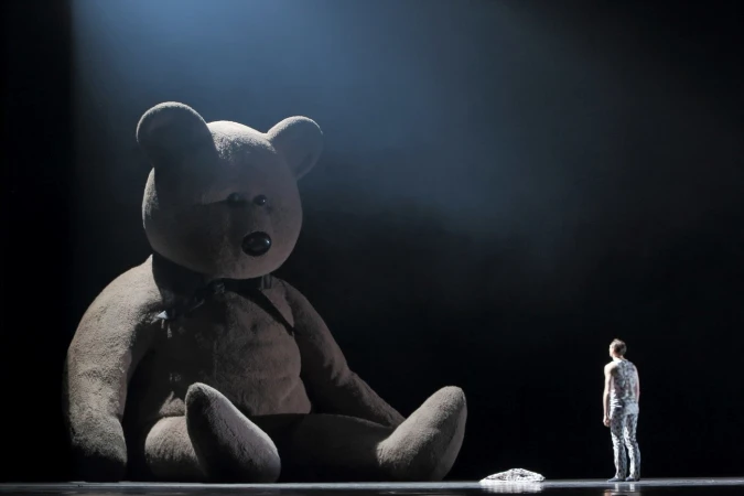 A giant teddy bear sits on a stage opposite a lone standing person under theatrical spotlighting in Sydney.