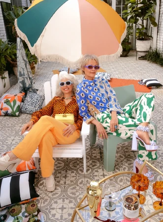 Two stylish senior women lounge in patterned outfits under a colorful umbrella on a patio with a tiled floor and cocktail accessories.