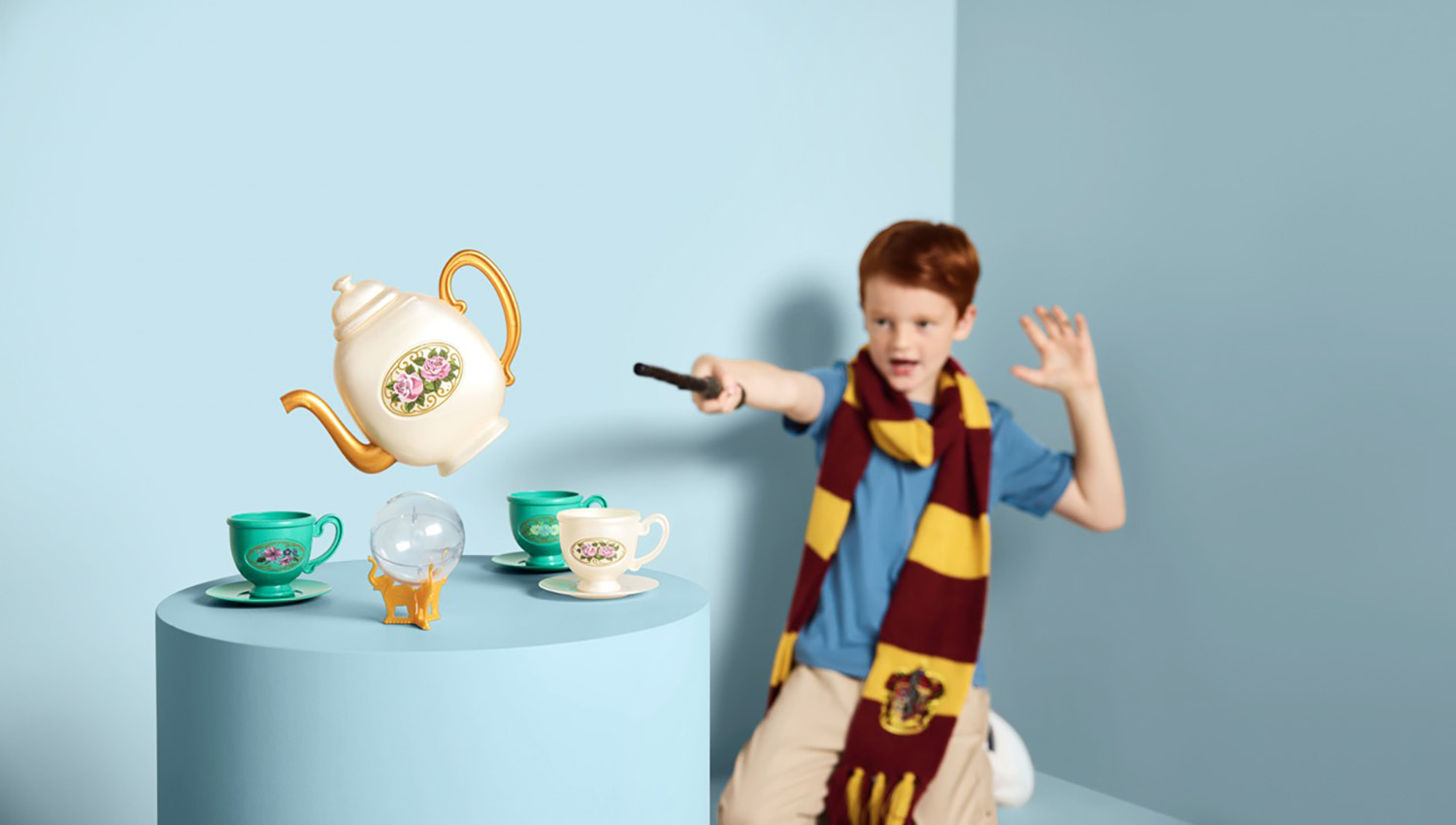 Young red-haired boy in striped scarf uses wand to levitate toy teapot, cups, and crystal ball against a blue studio background.