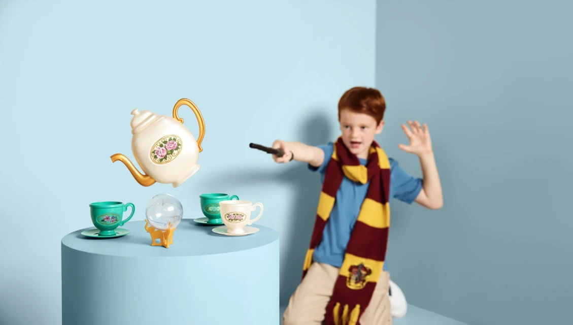 Young red-haired boy in striped scarf uses wand to levitate toy teapot, cups, and crystal ball against a blue studio background.