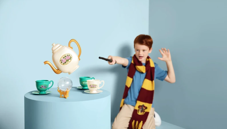 Young red-haired boy in striped scarf uses wand to levitate toy teapot, cups, and crystal ball against a blue studio background.