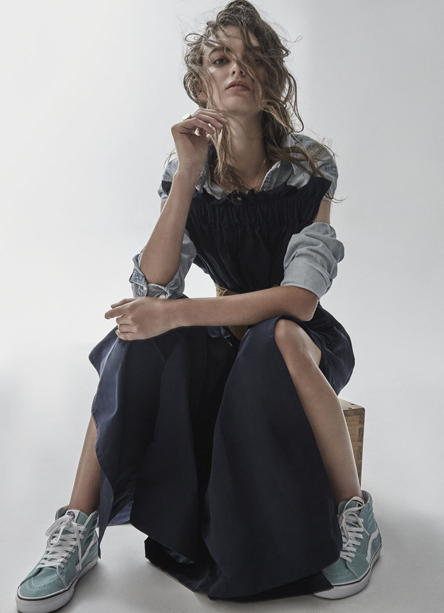 Adult female model wearing an open-shoulder blue dress with long denim sleeves, sitting on a wooden box in a studio, fashion editorial.