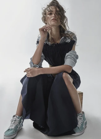 Model with tousled hair seated on a wooden crate wearing a black dress over a light shirt and teal sneakers against white