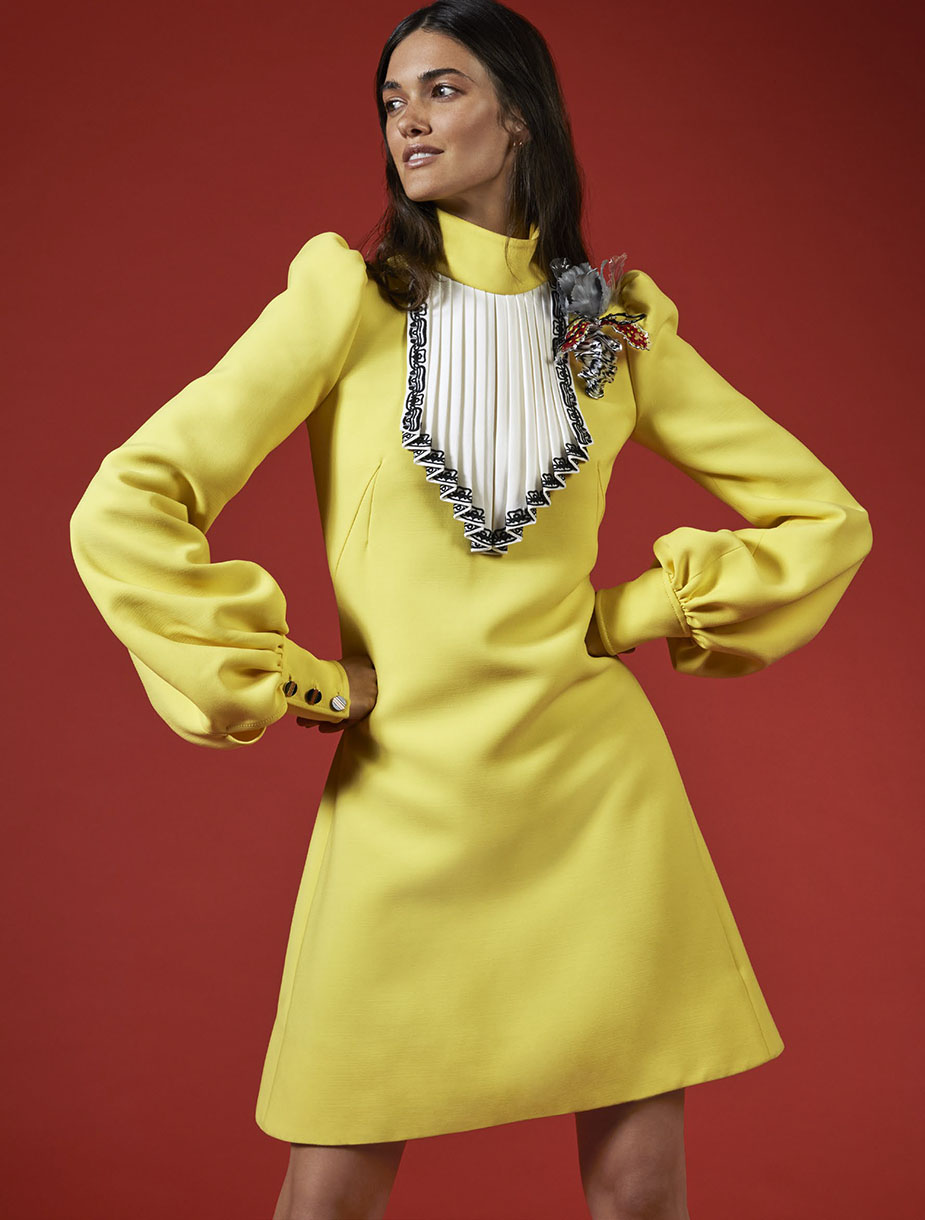 Elegant adult female in a yellow mini dress with puffy sleeves posing against a red studio backdrop; high fashion, studio shot.