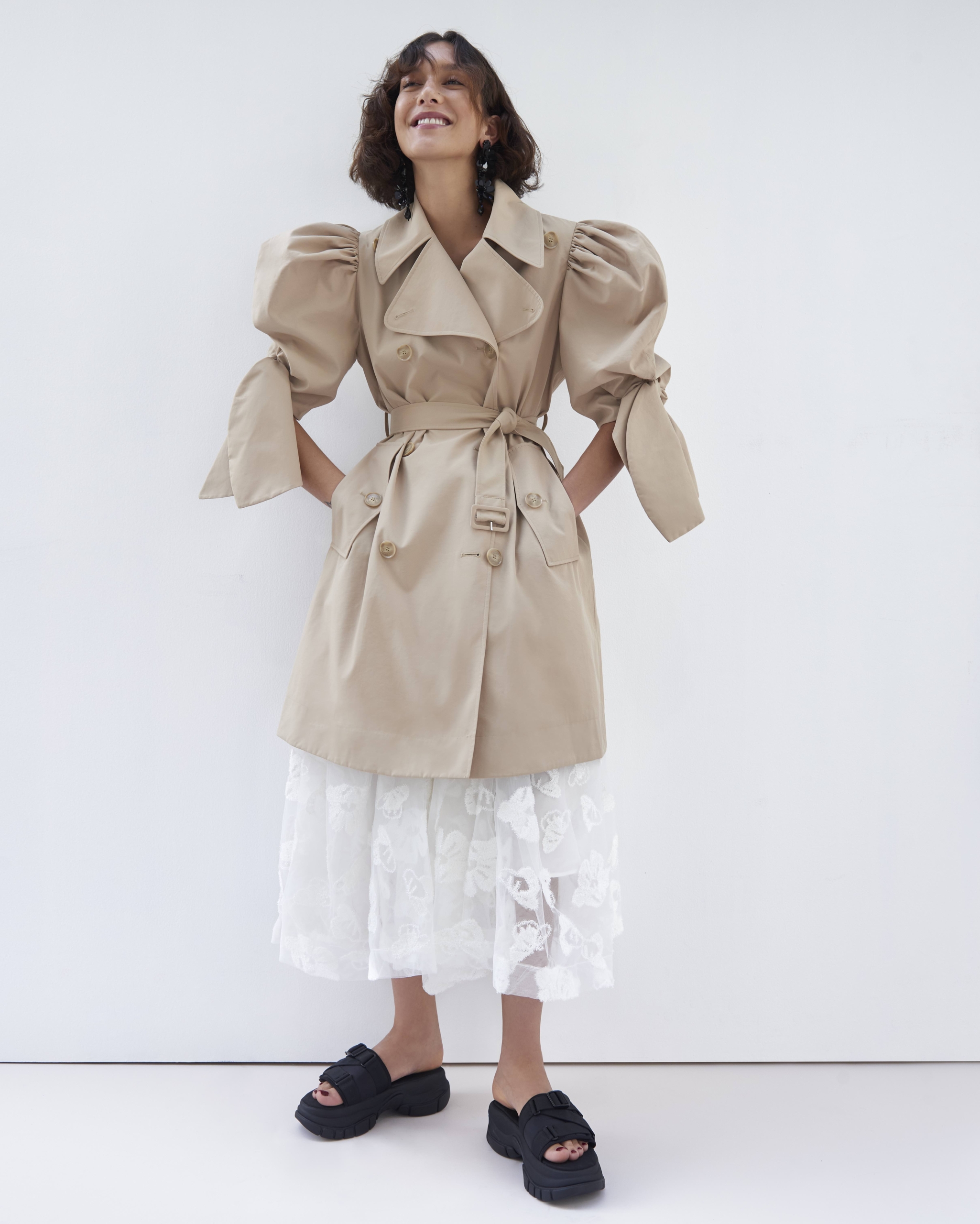 Smiling woman poses in a beige trench coat with balloon sleeves, a lace skirt, and black platform sandals against a white backdrop.