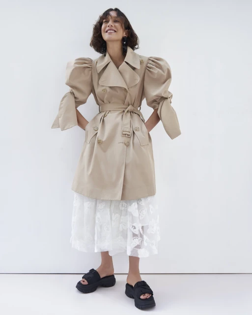 Smiling woman poses in a beige trench coat with balloon sleeves, a lace skirt, and black platform sandals against a white backdrop.