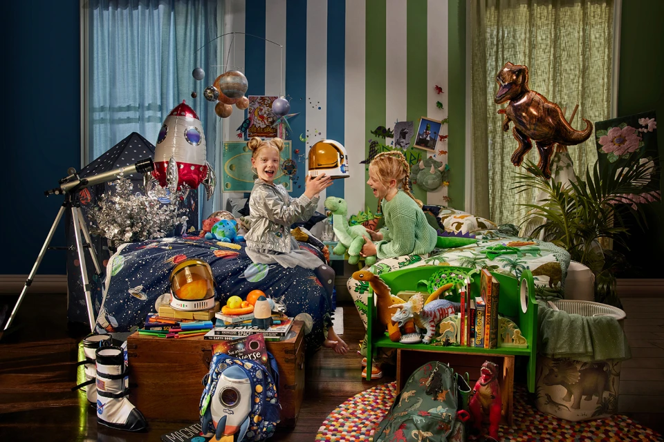 Two smiling young girls in a whimsical child's bedroom with outer space and dinosaur themes, surrounded by themed toys and decorations.