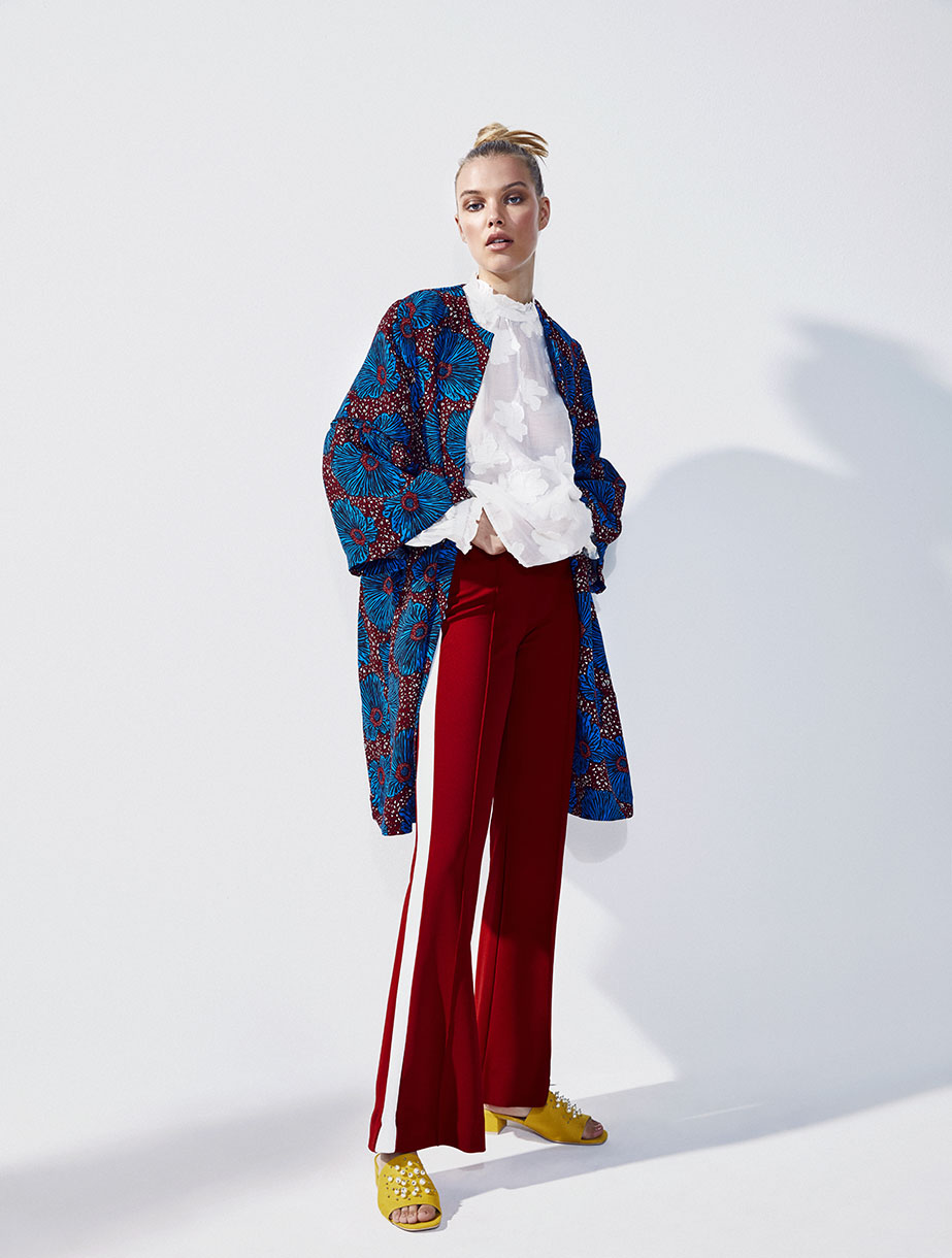Full-length shot of a blond-haired fashion model in a blue patterned coat, white blouse, red pants, and yellow sandals in a white photo studio.