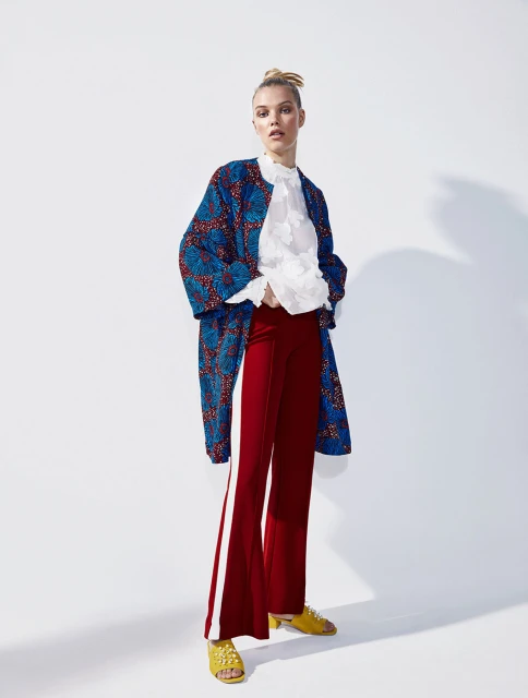 Full-length shot of a blond-haired fashion model in a blue patterned coat, white blouse, red pants, and yellow sandals in a white photo studio.
