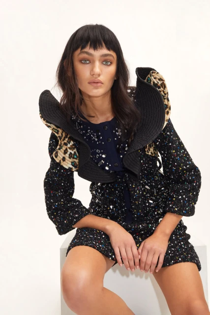 Brunette adult woman in a colorful, patterned mini dress with an oversized black and leopard print collar poses in a studio setting.