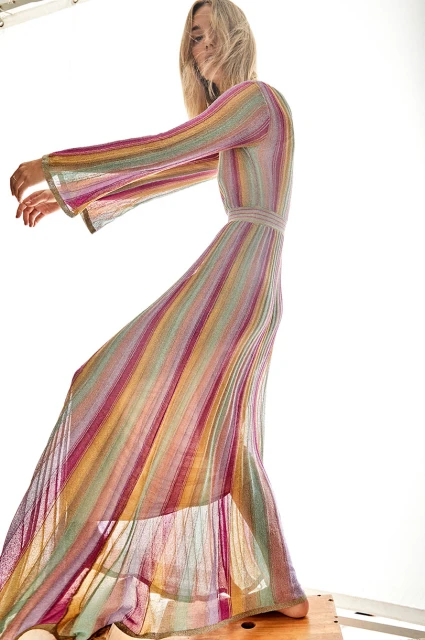 Fashion model in a full-length, colorful striped knit dress poses on a wooden platform in a bright, high-key lit studio setting.