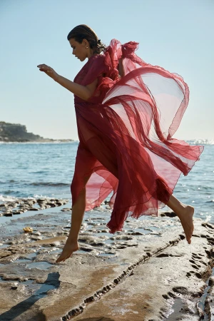 Woman in a sheer red dress leaping across rocky coastal shoreline with ocean in the background