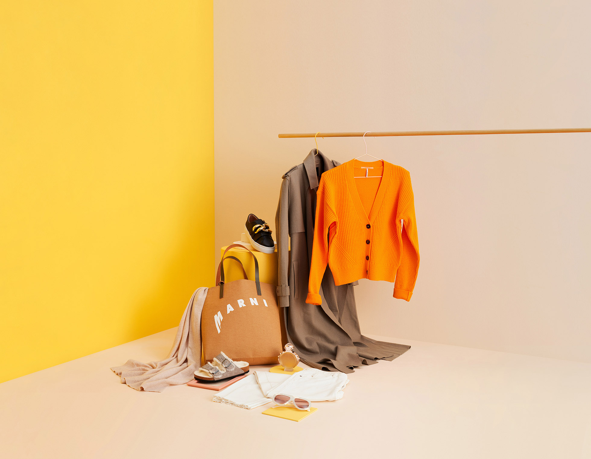 Fashion flat lay with an orange cardigan, tote bag, shoes, sunglasses, and other accessories against a yellow and beige studio backdrop.